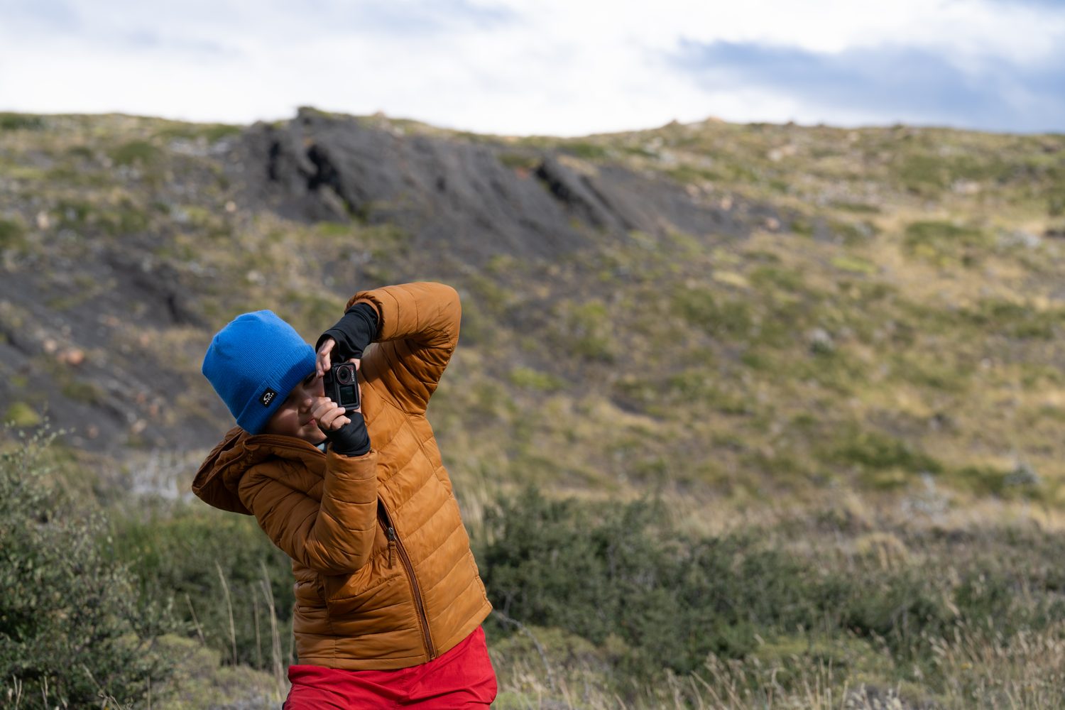 A young creator raising a small camera to their eye on a windswept hillside, standing off to one side of the frame.