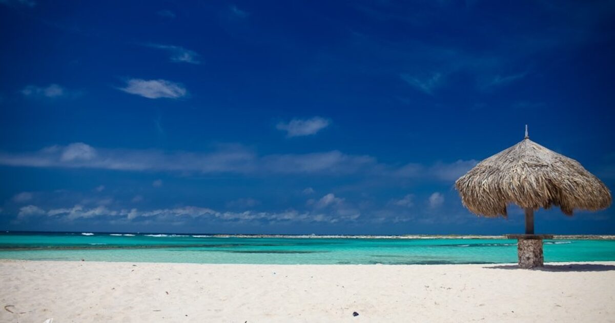 An empty white-sand beach with a single thatched palapa and a wide open sky, one landmark standing where a whole coastline could be